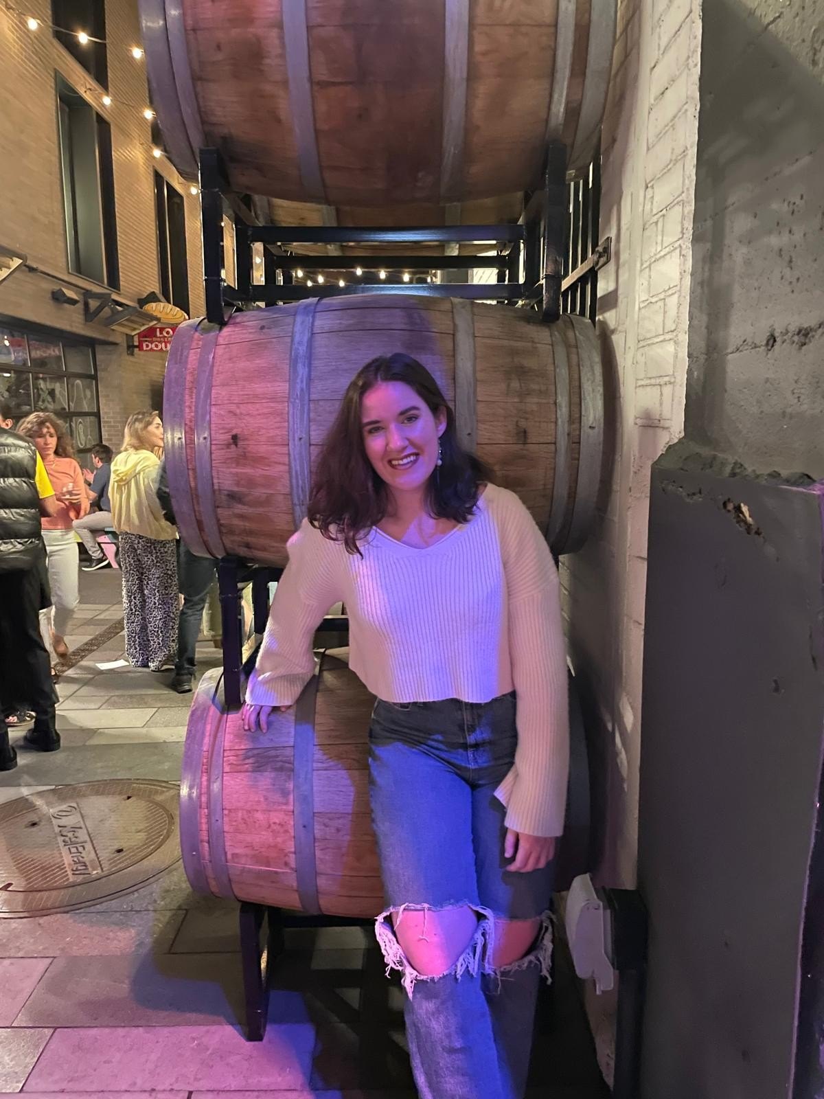 Caroline Moran sorri, apoiada em barris de madeira empilhados numa viela iluminada à noite