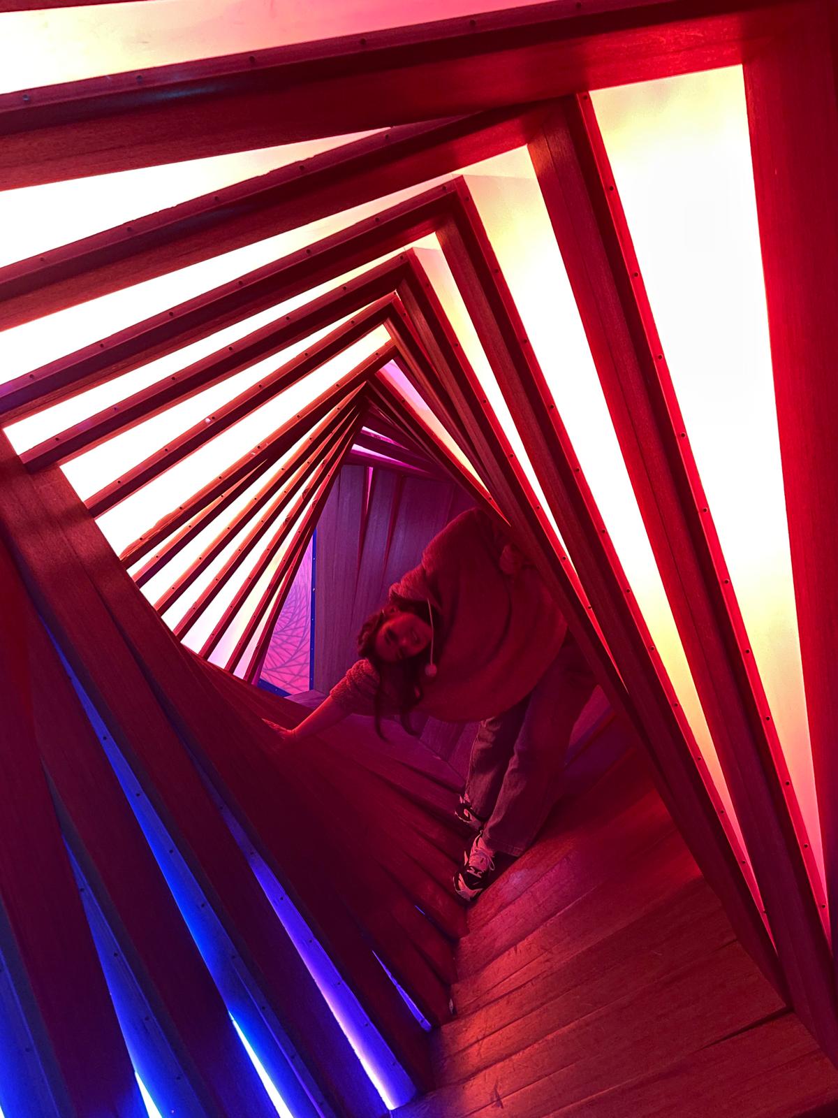 Uma pessoa se inclina dentro de um túnel de molduras de madeira iluminado de vermelho, rosa e azul