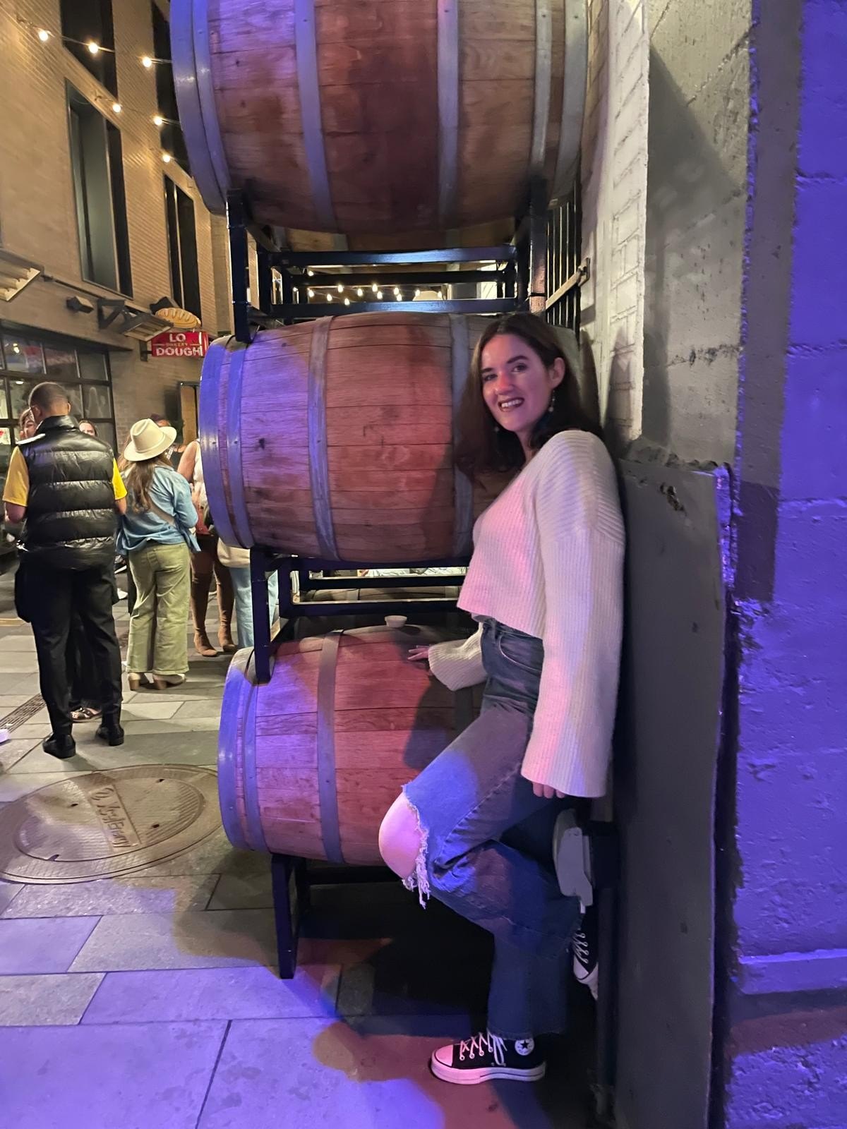 Caroline Moran encostada numa parede ao lado de barris de madeira, sob luz roxa, numa viela à noite