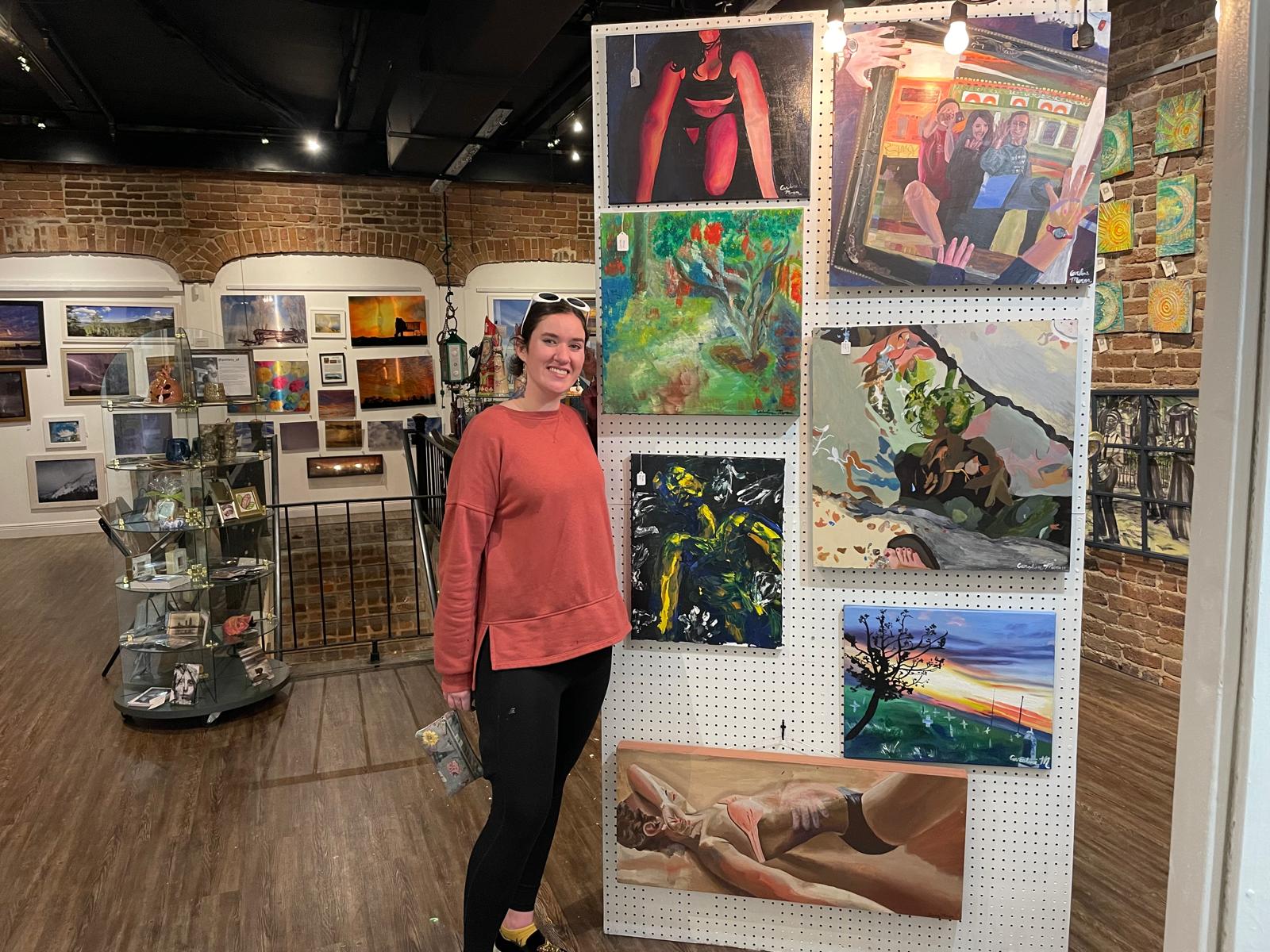 Caroline Moran sorri ao lado de um painel com sete de suas telas, numa galeria de paredes de tijolo