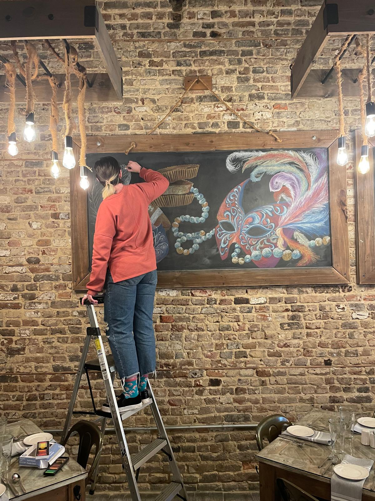 Pessoa de costas, em cima de uma escada, desenha com pastel um painel de giz com uma máscara de carnaval na parede de tijolos de um restaurante